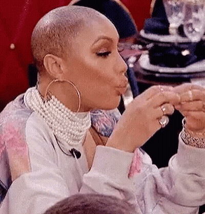 A Woman Wearing A Pearl Necklace And Hoop Earrings Is Sitting At A Table Eating A Piece Of Food . GIF