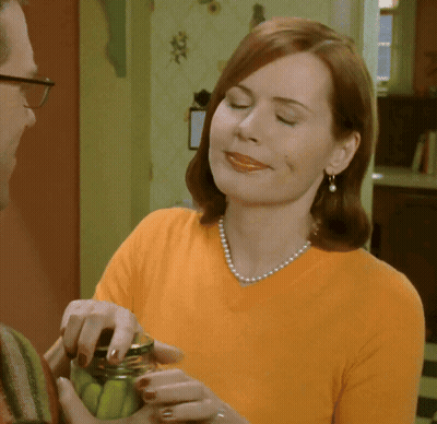 A Woman Wearing A Pearl Necklace Is Smiling While Holding A Jar Of Pickles GIF