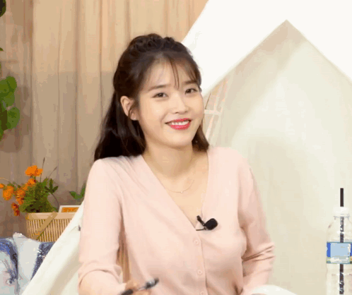 A Woman Wearing A Pink Cardigan And A Microphone Smiles GIF