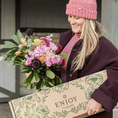 A Woman Wearing A Pink Hat Is Holding A Box Of Flowers That Says Enjoy Flowers GIF