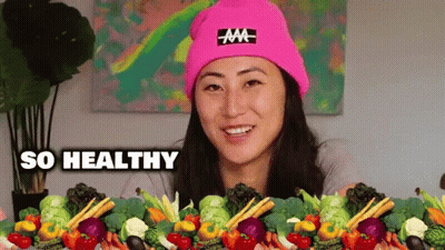 A Woman Wearing A Pink Hat Is Surrounded By Vegetables And The Words So Healthy Are Above Her . GIF