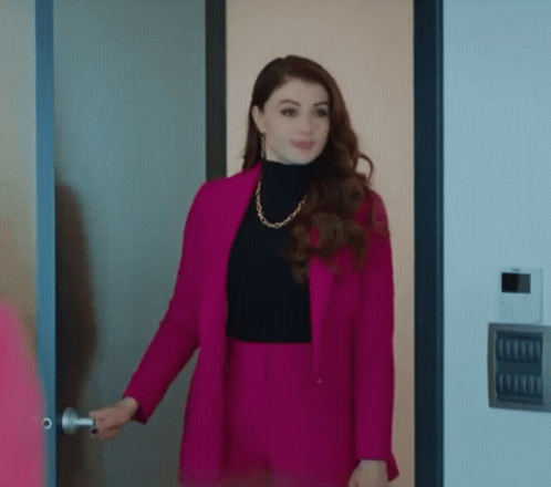 A Woman Wearing A Pink Jacket And Black Turtleneck Is Standing In A Doorway . GIF