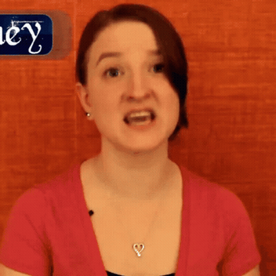 A Woman Wearing A Pink Shirt And A Heart Shaped Necklace Stands In Front Of A Sign That Says Hey GIF