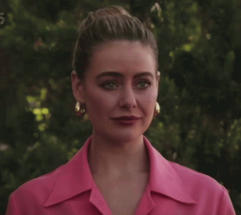 A Woman Wearing A Pink Shirt And Gold Hoop Earrings Stands In Front Of Trees GIF