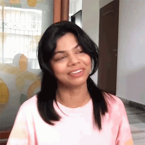 A Woman Wearing A Pink T-Shirt Is Smiling And Looking At The Camera GIF