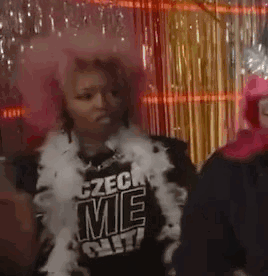 A Woman Wearing A Pink Wig And Feather Boa Is Standing Next To Another Woman Wearing A Black Shirt . GIF
