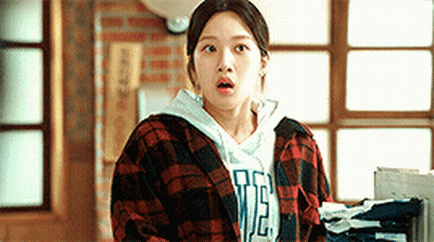 A Woman Wearing A Plaid Shirt And A Hoodie Is Standing In Front Of A Window . GIF