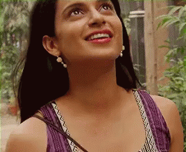 A Woman Wearing A Purple Tank Top And Earrings Is Smiling While Looking Up . GIF