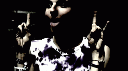 A Woman Wearing A Purple Tie Dye Shirt Is Giving A Peace Sign GIF