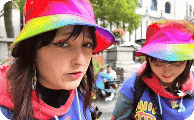 A Woman Wearing A Rainbow Hat Is Standing Next To Another Woman Wearing A Blue Shirt That Says ' Noodles ' GIF