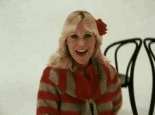 A Woman Wearing A Red And Gray Striped Sweater And A Red Flower In Her Hair Is Sitting In A Chair . GIF