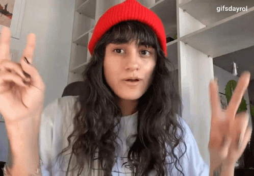 A Woman Wearing A Red Beanie And A White Shirt Is Making A Peace Sign GIF