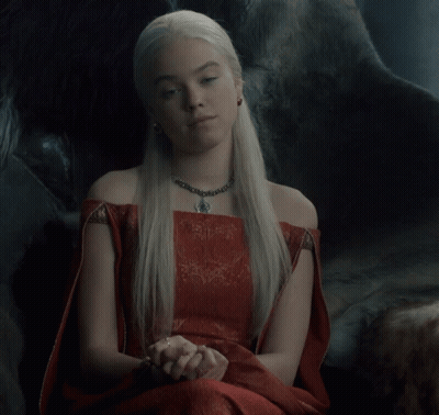 A Woman Wearing A Red Dress And A Necklace Sits In A Chair GIF