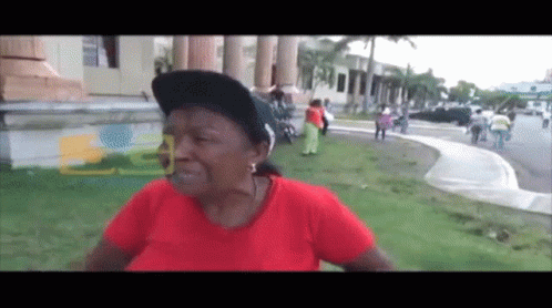 A Woman Wearing A Red Shirt And A Black Hat Stands In Front Of A Building With The Letter E On It GIF