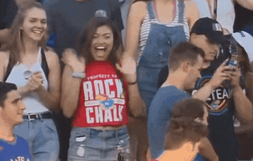 A Woman Wearing A Red Shirt That Says Rock Chalk Stands In A Crowd Of People GIF