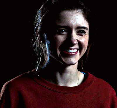 A Woman Wearing A Red Sweatshirt With A Pocket On The Front Smiles In The Dark GIF