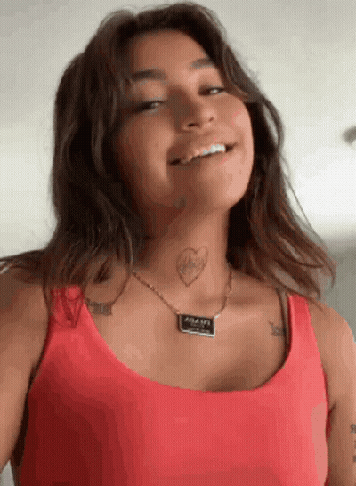 A Woman Wearing A Red Tank Top And A Necklace Has A Tattoo Of A Heart On Her Neck . GIF