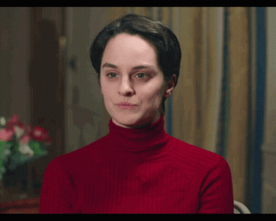 A Woman Wearing A Red Turtleneck Sweater Is Sitting In A Chair GIF