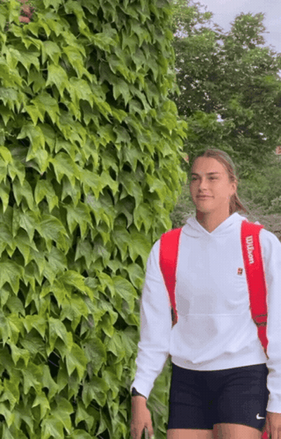 A Woman Wearing A Red Wilson Backpack Stands In Front Of A Lush Green Wall GIF