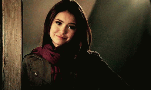 A Woman Wearing A Scarf And A Jacket Is Standing Next To A Wall GIF