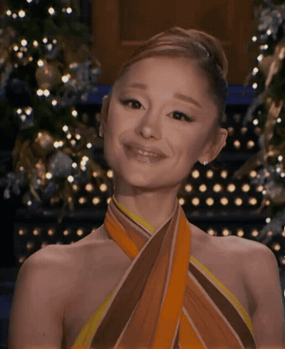 A Woman Wearing A Scarf Around Her Neck Is Smiling In Front Of A Christmas Tree . GIF