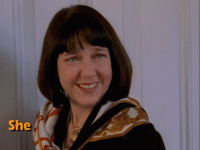 A Woman Wearing A Scarf Smiles In Front Of A Wall With The Word She Above Her GIF