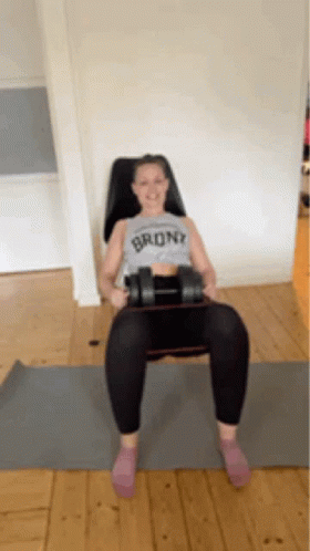 A Woman Wearing A Shirt That Says Bronx Is Sitting On A Yoga Mat Holding A Dumbbell GIF