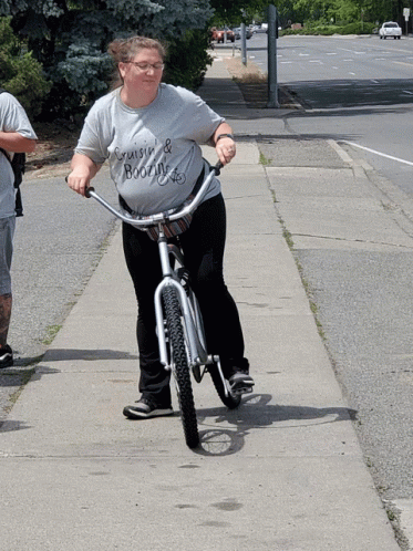 A Woman Wearing A Shirt That Says Cruise & Booze Is Riding A Bike Down A Sidewalk GIF