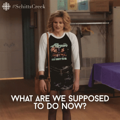 A Woman Wearing A Shirt That Says " What Are We Supposed To Do Now " Stands In A Room GIF