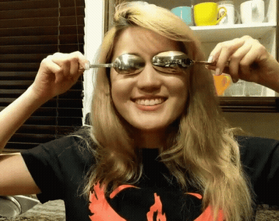 A Woman Wearing A Shirt With A Phoenix On It Is Holding A Spoon To Her Eye GIF