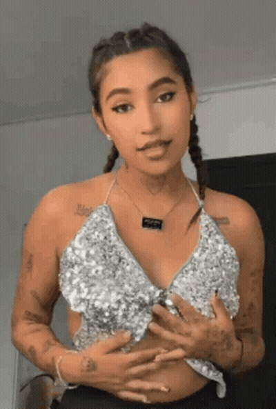 A Woman Wearing A Silver Top And A Necklace Is Standing In Front Of A Wall . GIF