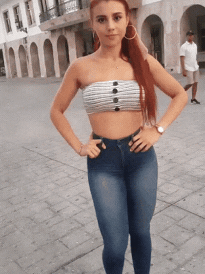 A Woman Wearing A Strapless Top And Jeans Stands On A Sidewalk With Her Hands On Her Hips GIF
