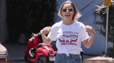 A Woman Wearing A Strauss & Co T-Shirt Is Dancing In Front Of A Red Scooter GIF