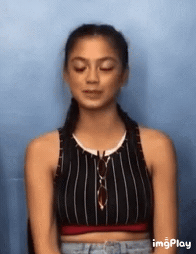 A Woman Wearing A Striped Crop Top And Jeans Is Sitting In Front Of A Blue Wall . GIF
