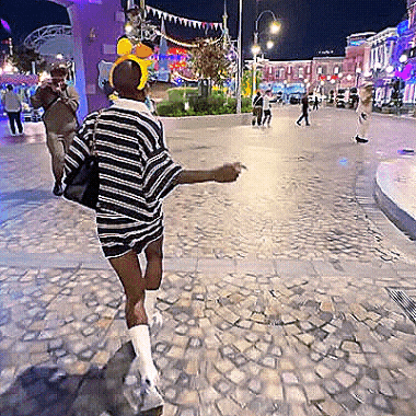 A Woman Wearing A Striped Shirt And Shorts Is Walking Down A Cobblestone Street GIF
