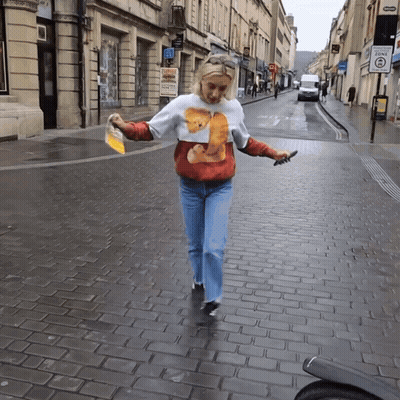 A Woman Wearing A Sweater With A Bear On It Is Walking Down A Brick Street GIF