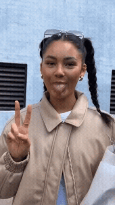 A Woman Wearing A Tan Jacket Is Sticking Her Tongue Out And Giving A Peace Sign GIF
