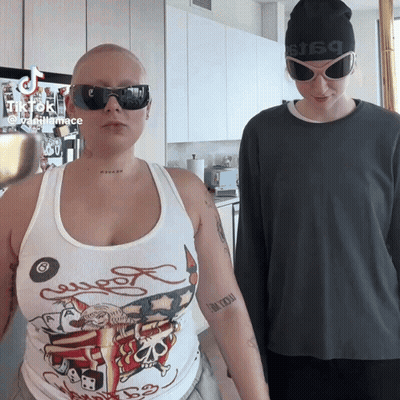A Woman Wearing A Tank Top That Says Ed Hardy Is Standing Next To A Man Wearing Sunglasses GIF