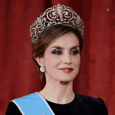 A Woman Wearing A Tiara And A Blue Sash Looks At The Camera GIF