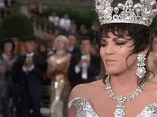 A Woman Wearing A Tiara And A Necklace Is Standing In Front Of A Crowd Of People . GIF