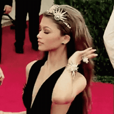 A Woman Wearing A Tiara And Bracelets Is Standing On A Red Carpet GIF