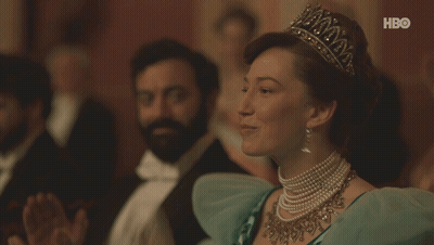 A Woman Wearing A Tiara And Pearls Sits In Front Of A Hbo Sign GIF