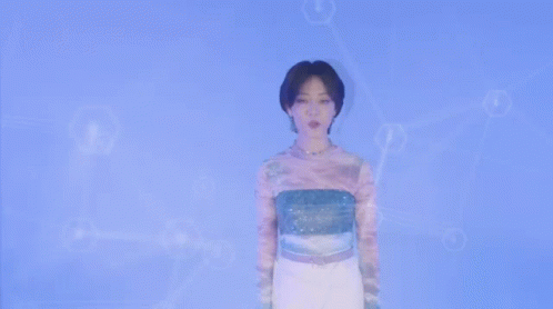 A Woman Wearing A Tie Dye Shirt And A Necklace Looks At The Camera . GIF