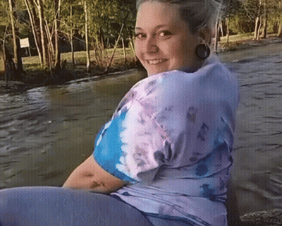 A Woman Wearing A Tie Dye Shirt Is Sitting On A Rock Near A River . GIF