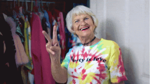 A Woman Wearing A Tie Dye Shirt That Says Party Is Love Gives A Peace Sign GIF
