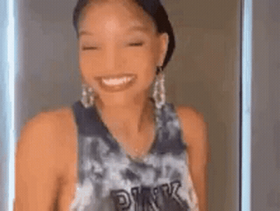 A Woman Wearing A Tie Dye Tank Top And Earrings Is Smiling . GIF