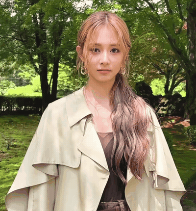 A Woman Wearing A Trench Coat And Earrings Is Standing In A Park GIF