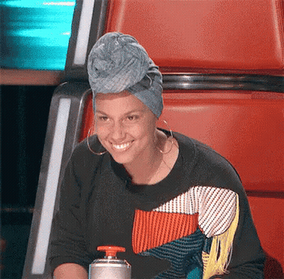 A Woman Wearing A Turban And Earrings Smiles While Sitting In A Chair GIF