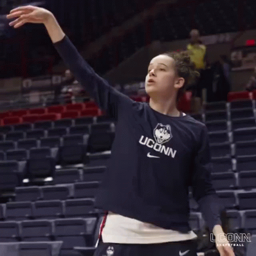 A Woman Wearing A Uconn Basketball Shirt Stretches Her Arms Out GIF