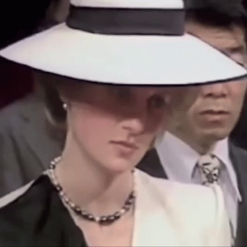 A Woman Wearing A White Hat And A Black Jacket Is Standing Next To A Man In A Suit . GIF